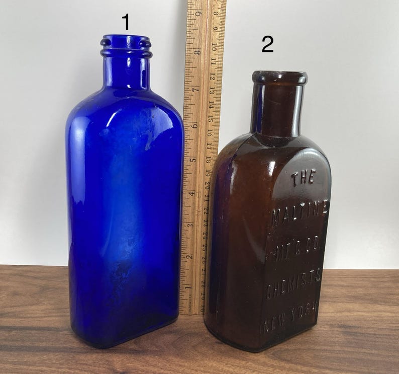 May include: Two antique glass bottles. One is a vibrant cobalt blue, the other is brown. The brown bottle has embossed text that reads "THE MALTINE MFG. CO. CHEMISTS NEW YORK". A wooden ruler is placed between the bottles.