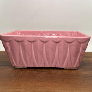May include: A rectangular pink ceramic planter featuring a repeating vertical scalloped pattern along the exterior walls. The vessel has a wide, open top and a smooth, glossy finish, showcasing a consistent pastel pink color throughout the entire piece.