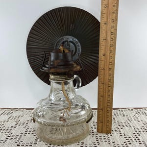 May include: A vintage clear glass oil lamp with a metal base and a brown metal reflector. The lamp has a handle and a wick. The wooden ruler next to the lamp shows it is approximately 7 inches tall.