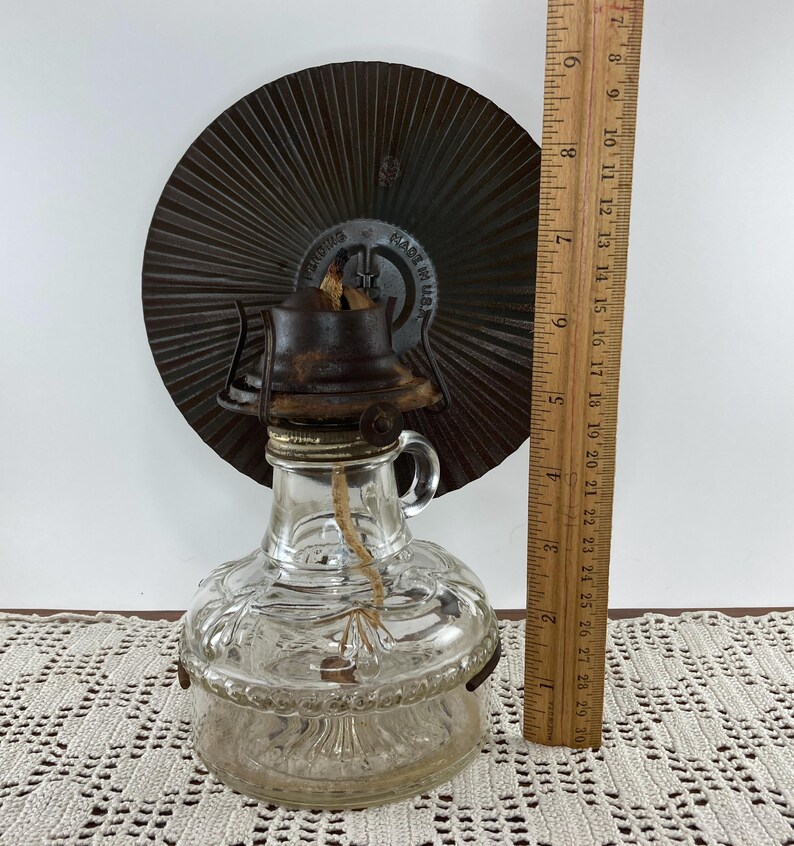 May include: A vintage clear glass oil lamp with a metal base and a brown metal reflector. The lamp has a handle and a wick. The wooden ruler next to the lamp shows it is approximately 7 inches tall.