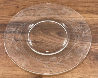 May include: A clear glass plate with etched floral and leaf designs. The plate has a circular shape with a slightly raised rim and a central recessed area. The etched design features flowers and leaves arranged around the plate's edge.