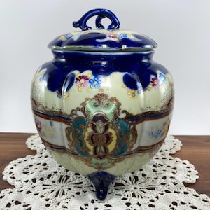 May include: A vintage ceramic jar with a lid. The jar is primarily cream-colored with a dark blue top and lid. It features floral and ornate gold accents, and rests on three small feet. The lid has a handle.