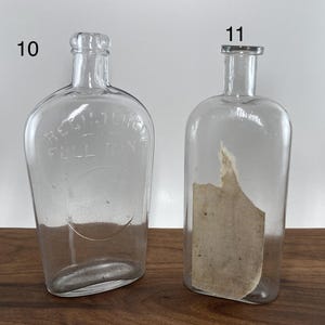 May include: Two clear glass bottles with rounded bodies and narrow necks. One bottle has the text "REGISTERED FULL PINT" embossed on the front. The other bottle has a partial label inside. Both bottles are empty.