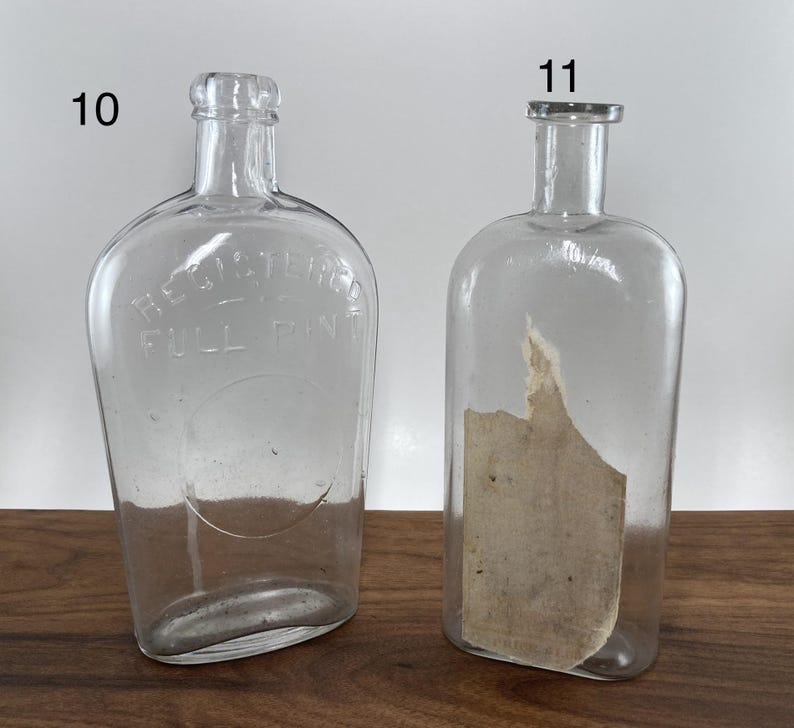 May include: Two clear glass bottles with rounded bodies and narrow necks. One bottle has the text "REGISTERED FULL PINT" embossed on the front. The other bottle has a partial label inside. Both bottles are empty.