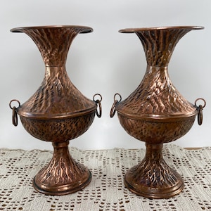 May include: Two antique copper vases with textured surfaces and ring handles. The vases have a flared top and a pedestal base. The copper has a rich, warm brown color. The vases are decorative and suitable for home decor.