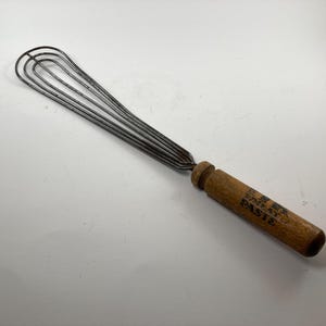 May include: A vintage wire whisk with a wooden handle. The handle is labeled "The Best Egg Beater Paste".