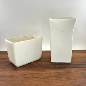 May include: Two off-white ceramic vessels with a smooth finish. One vessel is a short, wide rectangular container with rounded corners, while the other is a taller, slender rectangular vase with a slightly flared top opening and a flat base.