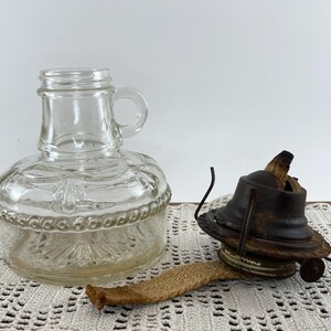 May include: A clear glass oil lamp base with a handle and decorative detailing, next to a vintage oil lamp burner. The burner is dark brown with a wick. The lamp is likely for home decor or ambiance.