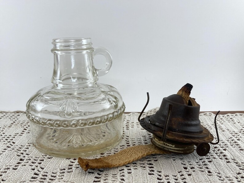 May include: A clear glass oil lamp base with a handle and decorative detailing, next to a vintage oil lamp burner. The burner is dark brown with a wick. The lamp is likely for home decor or ambiance.