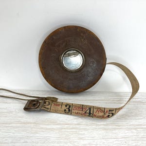 May include: Vintage brown leather measuring tape with a metal center. The tape extends from the circular case, displaying numbers and text. The tape measure is a classic tool for crafts, sewing, and construction, and is marked with the text "The Lufkin Rule Made in USA."