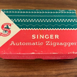 May include: A rectangular cardboard box for a Singer Automatic Zigzagger sewing machine attachment. The box features a red lower section, a dark green upper section with white stitch patterns, and a white corner with the red Singer logo.