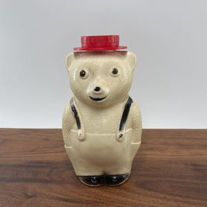 May include: A vintage bear-shaped honey dispenser. The bear is off-white with black accents on the eyes, nose, and feet. It wears a red hat and cream-colored overalls with black straps. The dispenser is made of ceramic.