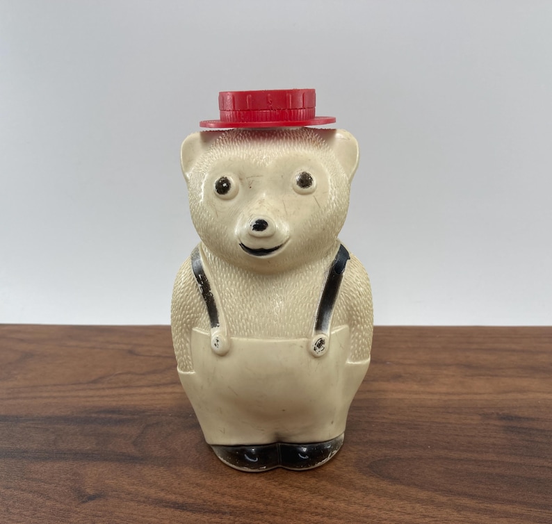 May include: A vintage bear-shaped honey dispenser. The bear is off-white with black accents on the eyes, nose, and feet. It wears a red hat and cream-colored overalls with black straps. The dispenser is made of ceramic.