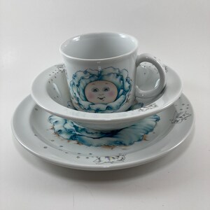 May include: A white porcelain children's dish set with a blue and white cabbage design. The set includes a bowl, a plate, and a mug. The cabbage has a smiling face.