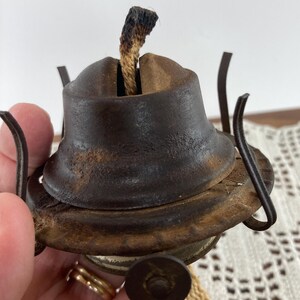 May include: Close-up of a vintage oil lamp burner. The metal burner is a dark, rusty brown with a wick protruding from the top. The lamp has a circular base and a small, round knob. The wick is a light brown color with a charred tip.