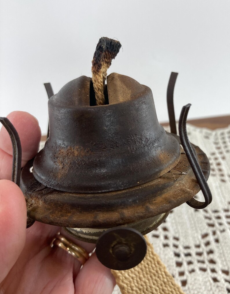 May include: Close-up of a vintage oil lamp burner. The metal burner is a dark, rusty brown with a wick protruding from the top. The lamp has a circular base and a small, round knob. The wick is a light brown color with a charred tip.