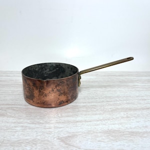 May include: A round copper saucepan with a weathered, dark patina on the exterior and a metallic interior. The pan features a long, straight brass handle attached to the side with two visible rivets, extending outward from the main body.