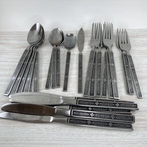 May include: A collection of stainless steel silverware, including spoons, forks, knives, and a butter knife. The handles feature an ornate, engraved design. The silverware is arranged on a light-colored surface.