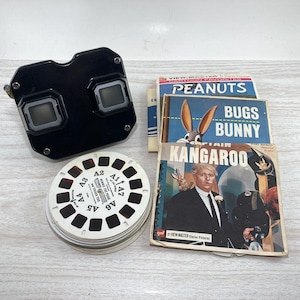 May include: A vintage black View-Master viewer with a stack of image reels and several View-Master packets. The packets feature the titles "Peanuts", "Bugs Bunny", and "Kangaroo". The image reels are white with black and red details.