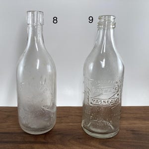 May include: Two clear glass antique bottles. One bottle has a smooth surface, while the other has embossed text that reads "SOLUTION CITRATE MAGNESIA". Both bottles have a narrow neck and a rounded base. The bottles are displayed on a wooden surface.