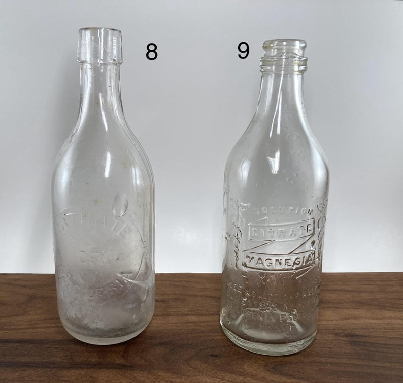 May include: Two clear glass antique bottles. One bottle has a smooth surface, while the other has embossed text that reads "SOLUTION CITRATE MAGNESIA". Both bottles have a narrow neck and a rounded base. The bottles are displayed on a wooden surface.