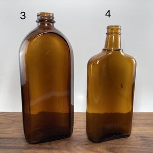 May include: Two amber-colored glass bottles. The bottle on the left has a rounded top and a rectangular base. The bottle on the right has a rectangular shape with a narrow neck. Both bottles are empty and sit on a wooden surface.