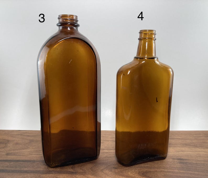 May include: Two amber-colored glass bottles. The bottle on the left has a rounded top and a rectangular base. The bottle on the right has a rectangular shape with a narrow neck. Both bottles are empty and sit on a wooden surface.