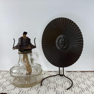May include: A clear glass oil lamp with a metal top and handle, next to a dark brown metal fan on a stand. The lamp has a wick and is decorated with embossed patterns. The fan has a radial design.