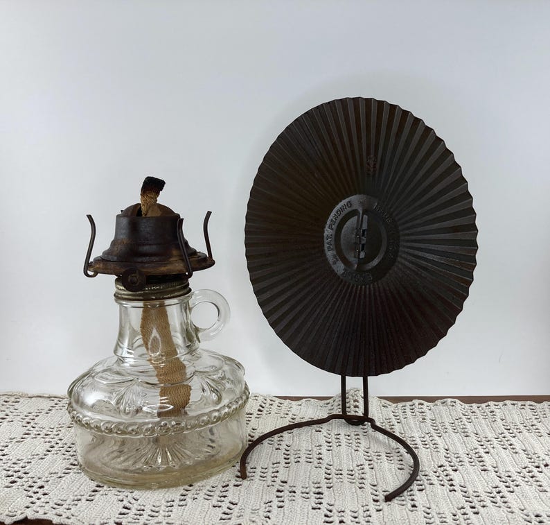 May include: A clear glass oil lamp with a metal top and handle, next to a dark brown metal fan on a stand. The lamp has a wick and is decorated with embossed patterns. The fan has a radial design.