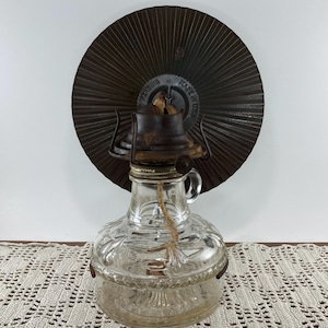 May include: A vintage clear glass oil lamp with a decorative embossed pattern. The lamp has a metal top and a wick. A large, circular, brown metal reflector is behind the lamp. The reflector has a radial design and the text "MADE IN USA".