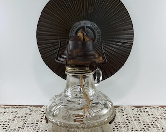 May include: A vintage clear glass oil lamp with a decorative embossed pattern. The lamp has a metal top and a wick. A large, circular, brown metal reflector is behind the lamp. The reflector has a radial design and the text "MADE IN USA".