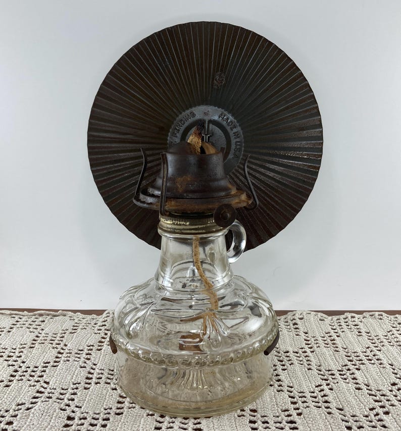 May include: A vintage clear glass oil lamp with a decorative embossed pattern. The lamp has a metal top and a wick. A large, circular, brown metal reflector is behind the lamp. The reflector has a radial design and the text "MADE IN USA".