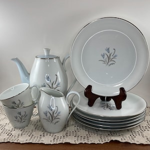 May include: A vintage white porcelain tea set with a light blue floral design and silver trim. The set includes a teapot, creamer, sugar bowl, cups, and plates. The plates are stacked and displayed on a wooden stand.