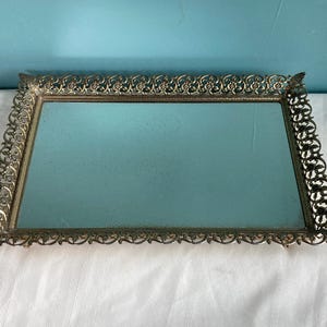 May include: Rectangular antique vanity tray with a mirrored surface and ornate filigree metal frame. The frame has a gold tone with floral and scroll detailing. The tray is approximately 12 inches long and 8 inches wide.