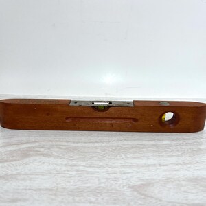 May include: A rectangular wooden spirit level featuring a smooth, medium-brown finish. The tool includes a central horizontal glass vial filled with yellow liquid and a circular vertical vial on the right side, both set into the wood frame.