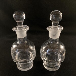 May include: Two clear glass bottles with stoppers. The bottles have a round body and a textured surface.