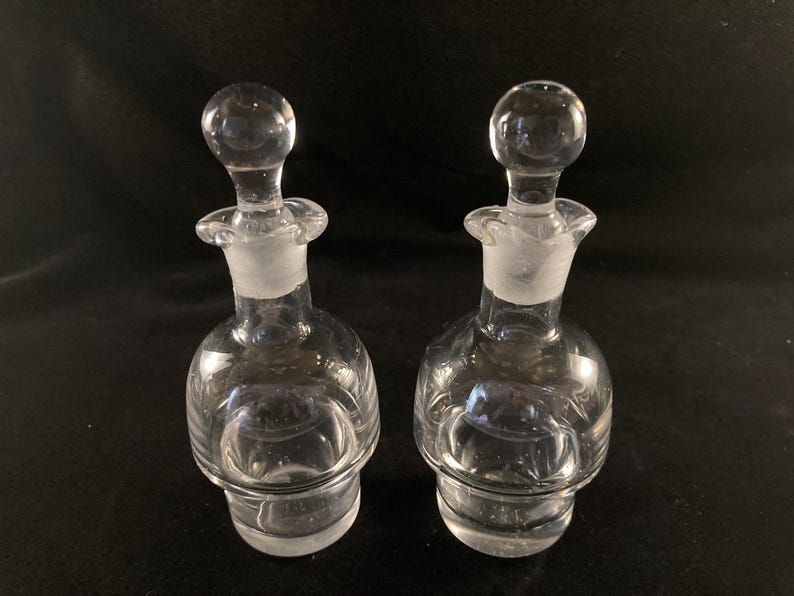 May include: Two clear glass bottles with stoppers. The bottles have a round body and a textured surface.