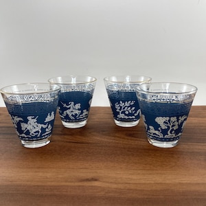 May include: Four clear drinking glasses with a blue and white design. The glasses feature a blue band with a white illustration of a rider on a horse, trees, and other decorative elements. The glasses are on a wooden surface.