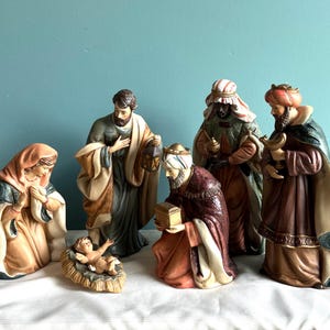 May include: A nativity set featuring six figurines: a kneeling figure in a peach and blue robe, a standing figure in a tan and blue robe holding a lantern, a baby in a manger, and three standing figures in ornate robes holding a chest and vessels.