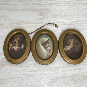 May include: Antique triptych featuring three oval frames linked by a chain. Each frame has a gold-toned metal finish and contains a sepia-toned portrait. The portraits depict cherubs and a mother with a baby, evoking a vintage aesthetic.