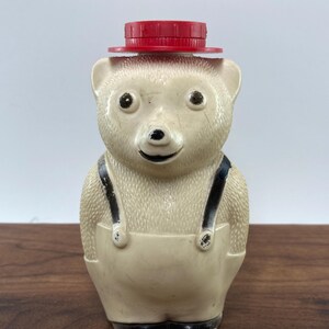 May include: A vintage bear-shaped container with a red lid, black suspenders, and black shoes. The bear is off-white with a smiling face. The container is likely made of plastic and is a collectible item.