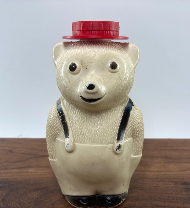 May include: A vintage bear-shaped container with a red lid, black suspenders, and black shoes. The bear is off-white with a smiling face. The container is likely made of plastic and is a collectible item.