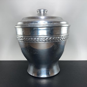 May include: A silver-colored metal container with a lid. The container has a rounded body with a decorative band of braided design around the middle. The lid has a small, rounded handle. The container is likely for storage.