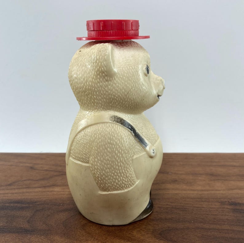 May include: A vintage honey bear-shaped bottle with a red cap. The bear is wearing overalls with black straps. The bottle is beige with black accents on the feet.