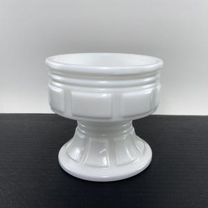 May include: A white, pedestal-style bowl with a wide, open top. The bowl has a decorative, geometric design around the middle and a flared base. The bowl is made of a glossy, opaque material and sits on a dark surface.