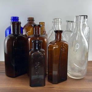 May include: A collection of antique glass bottles in various shapes and sizes. The bottles are in shades of brown, blue, and clear glass. Some bottles have embossed text. The bottles are arranged on a wooden surface.