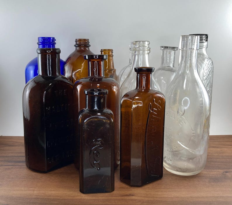 May include: A collection of antique glass bottles in various shapes and sizes. The bottles are in shades of brown, blue, and clear glass. Some bottles have embossed text. The bottles are arranged on a wooden surface.