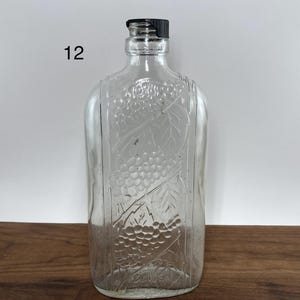 May include: Clear glass bottle with a black cap. The bottle has a rectangular shape with rounded edges and a grape design embossed on the front. The number "12" is printed on the left side of the bottle.