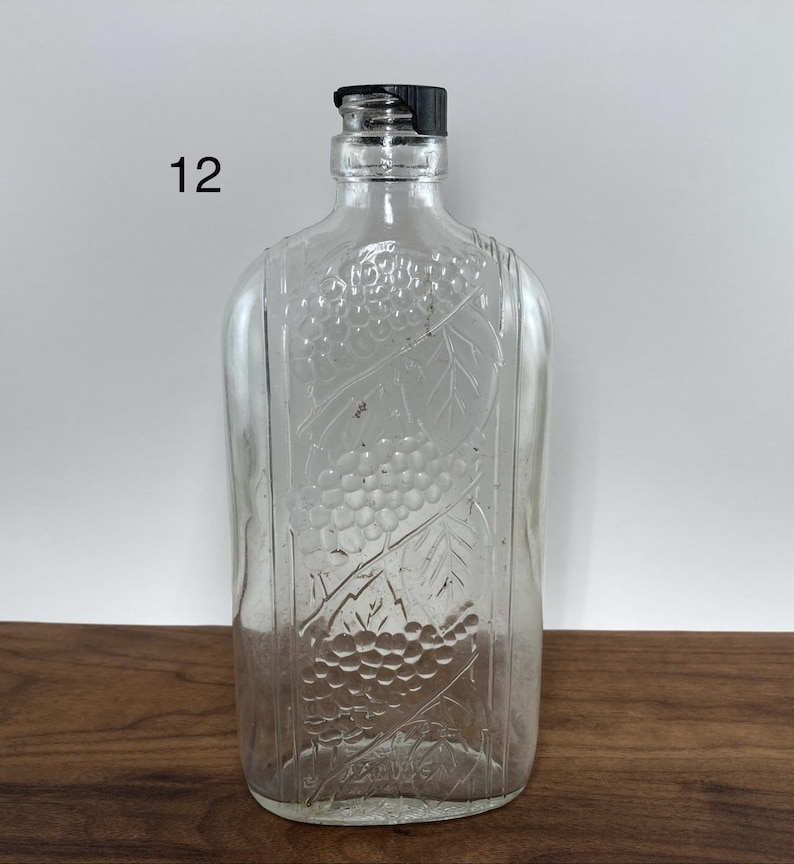 May include: Clear glass bottle with a black cap. The bottle has a rectangular shape with rounded edges and a grape design embossed on the front. The number "12" is printed on the left side of the bottle.