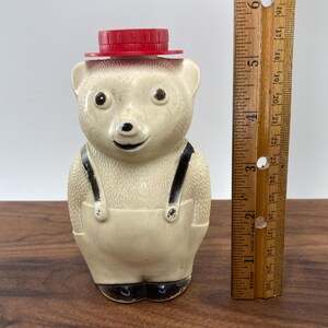 May include: A vintage bear-shaped honey bottle with a red cap. The bear is off-white with black accents on the eyes, nose, and suspenders. The bottle is approximately 5 inches tall, as measured by the wooden ruler.